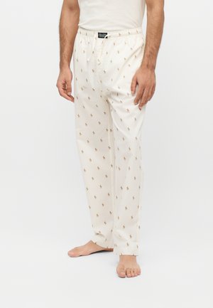 Homme portant un pantalon de pyjama blanc avec un petit motif imprimé de cheval et cavalier, debout pieds nus devant un fond uni.
