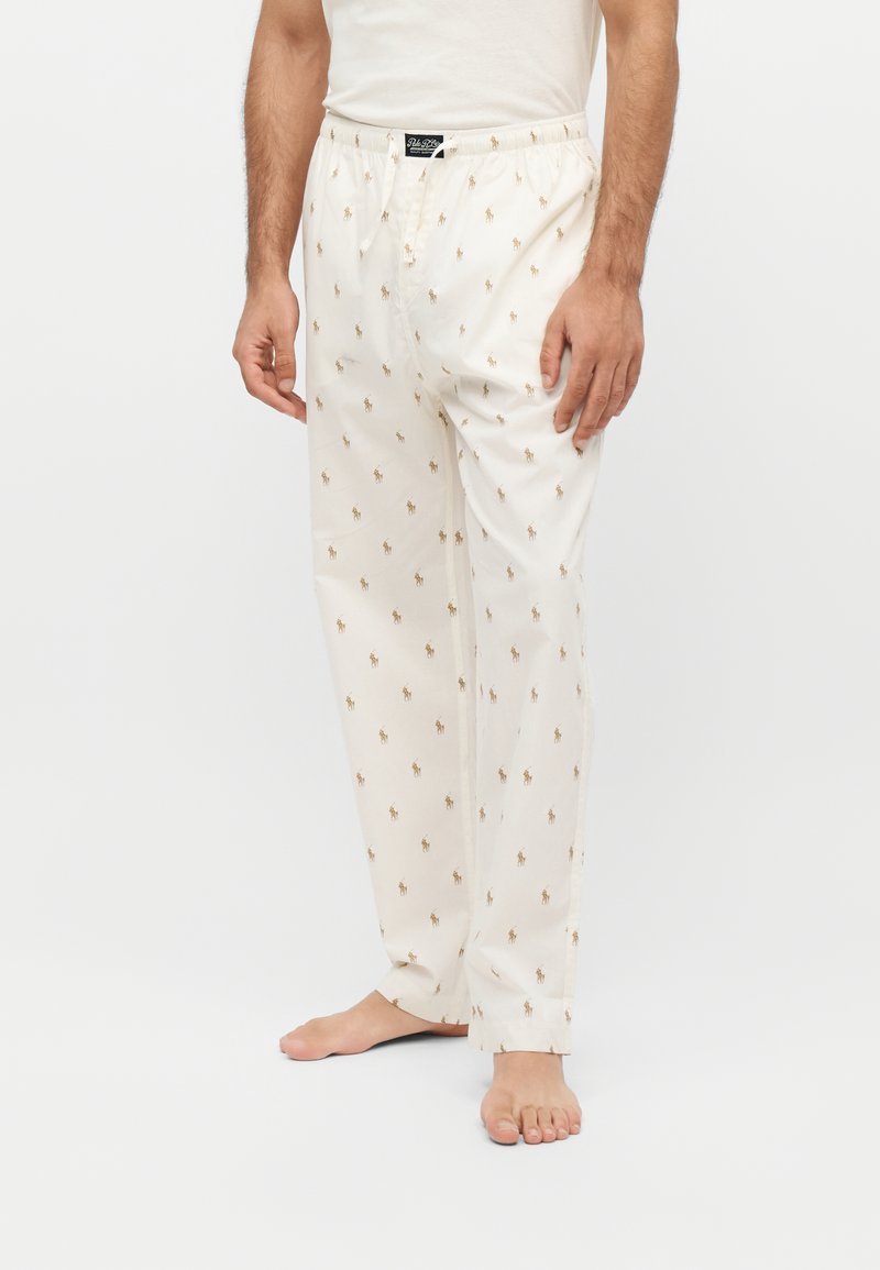 Homme portant un pantalon de pyjama blanc avec un petit motif imprimé de cheval et cavalier, debout pieds nus devant un fond uni.