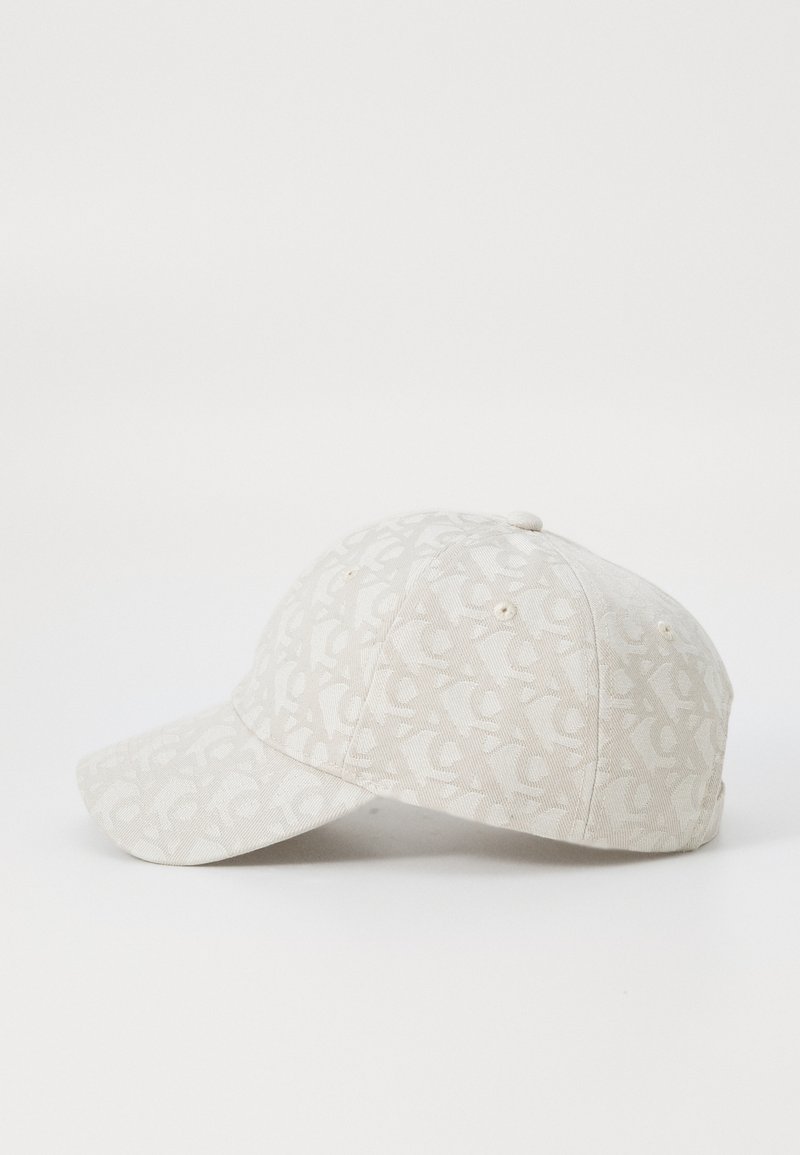 Gorra de color beige claro hecha de tela, con un sutil patrón de logotipos, diseño estructurado, visera curvada y acentos metálicos.