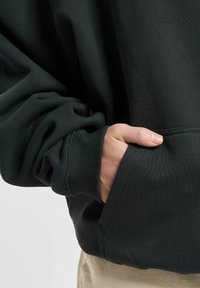 Mørkegrønn hettegenser med teksturert stoff, med en stor lomme foran og ribbede mansjetter. Materialet ser mykt og tykt ut.