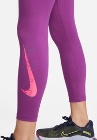 Fioletowe legginsy sportowe z elastycznego materiału, z wyraźnym różowym logo Nike oraz szarymi literami na dolnej części nogi.
