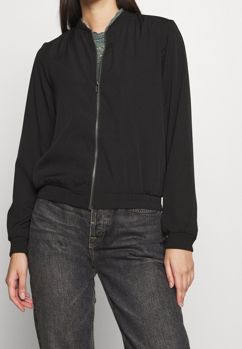 Czarna kurtka bomberka z zapięciem na zamek, elastycznymi mankietami i dołem. Gładki materiał z lekkim połyskiem. Noszona na fakturowanym zielonym topie.