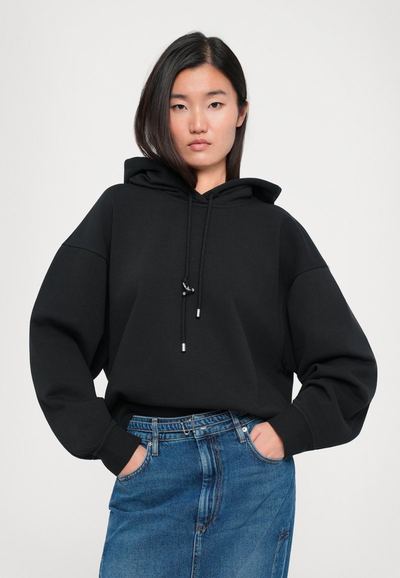 Haut de survêtement noir surdimensionné avec cordons de serrage et embouts en métal argenté, associé à une jupe en denim taille haute. Le tissu semble lisse et doux.