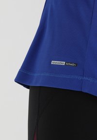 Blå aktivkläder skjorta med texturerat tyg, med en svart etikett märkt "AdvancedSkin ActiveDry" vid fållen, i kombination med svarta leggings.