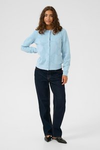 Hellblauer Cardigan aus weichem Material mit Knopfschluss und strukturiertem Muster, kombiniert mit dunkelblauen Denim-Hosen und schwarzen spitzen Schuhen.