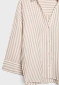 Beige-weiße, vertikal gestreifte Hemdjacke mit Knöpfen, langen Ärmeln und Bündchenärmeln, auf einer ebenen Fläche dargestellt.