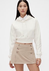 Hvit cropped hettegenser med snor og preget logo, kombinert med en beige miniskjørt med knappedetaljer foran.