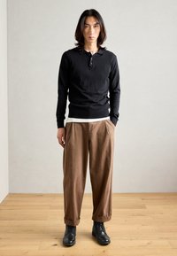 Schwarzes Langarm-Polo mit drei Knöpfen, kombiniert mit braunen gefalteten Hosen und schwarzen Schnürstiefeln. Glatte Textur und entspannter Schnitt.