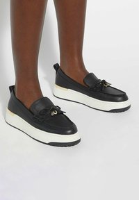 Black leather slip-on shoes with a textured upper, white sole, and gold accent at the heel. Features a lace bow and minimalist design.