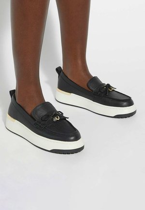 Black leather slip-on shoes with a textured upper, white sole, and gold accent at the heel. Features a lace bow and minimalist design.