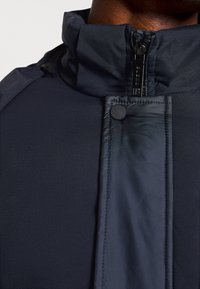 Tamno plava jakna s visokim ovratnikom, koja ima zatvaranje na zip i okrugli gumb na dugme. Glatka tekstura s kontrastnim detaljima od tkanine.