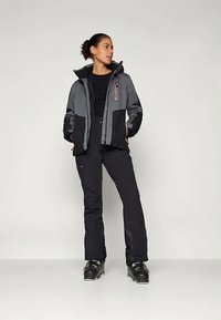 Femeie stand cu bocanci de schi negri, pantaloni negri, top negru și jachetă de schi gri și neagră, privind spre stânga pe un fundal simplu.