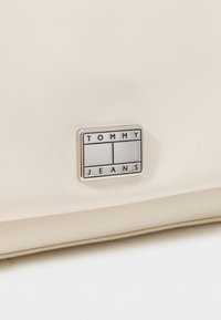 Bolso de cuero sintético blanco con una placa rectangular plateada con "TOMMY" y "JEANS" en relieve en negro. Textura suave con un diseño minimalista.
