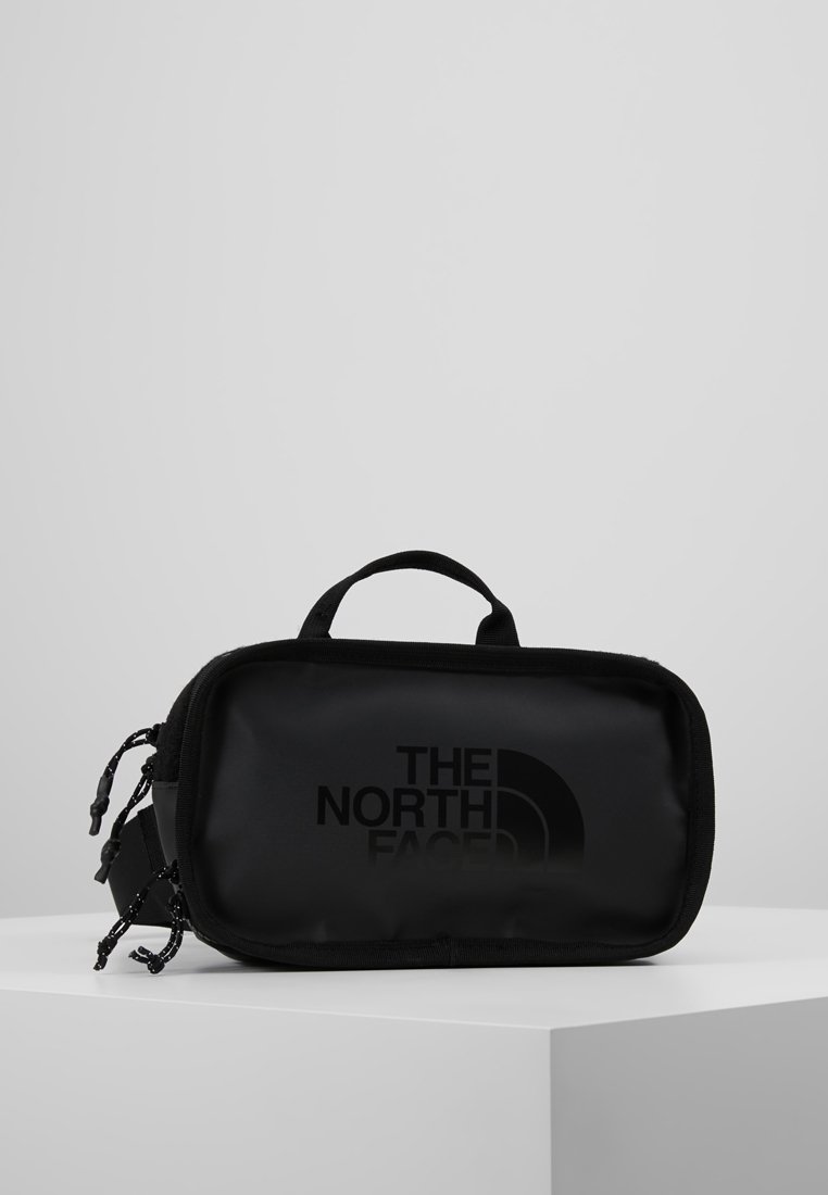 Trousse rectangulaire noire North Face avec fermeture éclair et poignée, placée sur un socle blanc sur un fond gris uni.