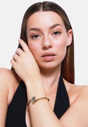 Bracciale in metallo con un design morbido e curvo, caratterizzato da finiture in oro e argento a contrasto. Indossato al polso, mette in mostra una texture elegante.