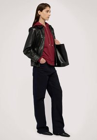 Černá kožená bunda přes vínovou mikinu s kapucí, v kombinaci s tmavě modrými širokými kalhotami a černými špičatými botami. Modelka drží černou tašku typu tote.