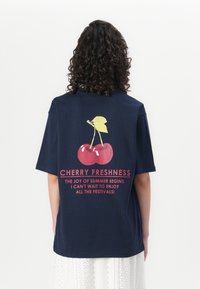 T-shirt overdimensionato blu navy con grafica di ciliegie e testo rosso. Il tessuto in cotone ha una texture liscia e una vestibilità comoda.