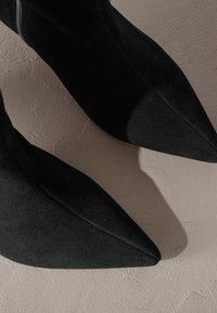 Czarne zamszowe botki na szpilce z ostrym noskiem o eleganckim designie i gładkiej teksturze, z bocznym zamkiem błyskawicznym. Umieszczone na jasnej, teksturowanej powierzchni.