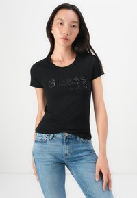 Žena u crnoj uskoj majici s natpisom "Guess est. 1981" i svijetloplavim trapericama, koja stoji ispred obične bijele pozadine.