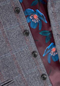 Grijze visgraatblazer met rode streepjes en zwarte knopen over een bordeaux shirt met grote blauwe bloemenprints.