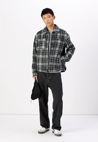 Grey and black plaid jacket with a collar, two front pockets, worn over a grey shirt and paired with black pants and grey sneakers.