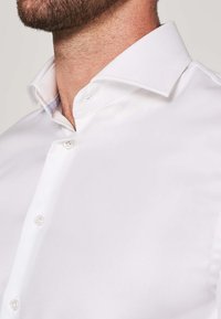 Weißes Hemd mit spitzem Kragen und Knopfverschluss, aus glattem Stoff. Sichtbare Nähte und Kragendetails.