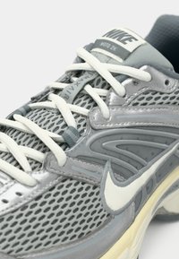 Zapatillas Nike con parte superior de malla gris, detalles en blanco y suela de goma. Incluyen cordones, marca visible y superposiciones texturizadas.