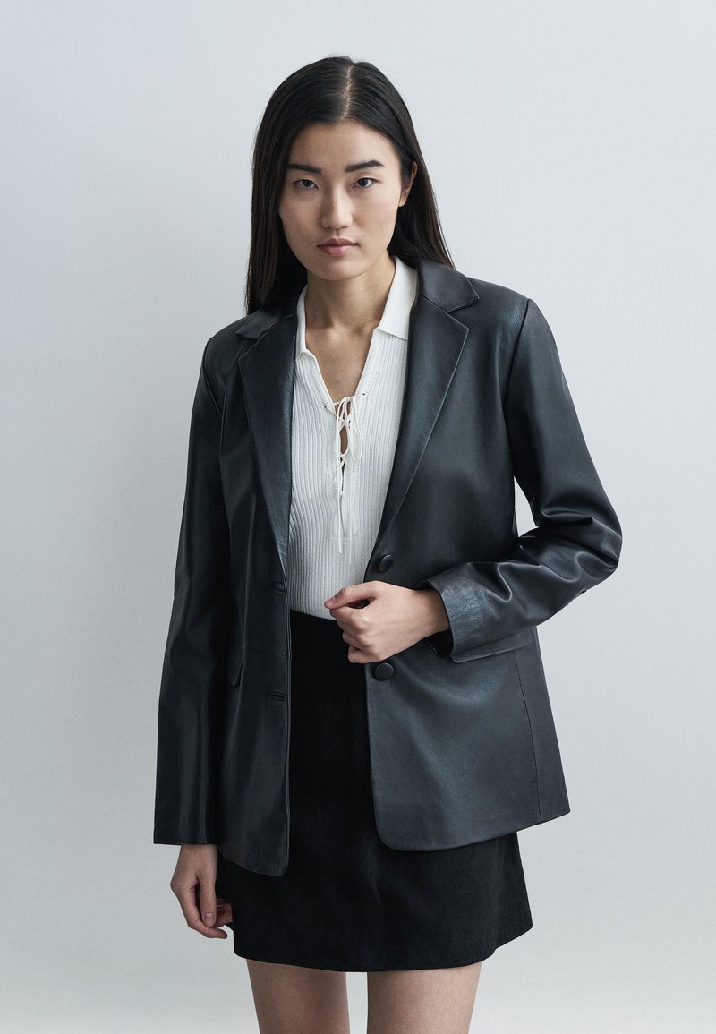 Black leather blazer with a tailored fit, notched lapels, two front buttons, and a smooth texture, paired with a white textured blouse.