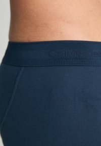 Tumedatest ribastest bokserpüksid pehme elastse vööga, millel on kirjaga "Calvin Klein" reljeefne logo. Sile tekstuur ja liibuv disain.