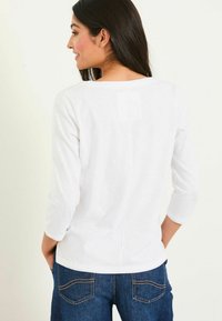 Weißes Oberteil mit drei Viertelärmeln und rundem Ausschnitt, mit einer mittigen Nahtdetail und weicher, glatter Textur. Getragen mit blauen Jeans.