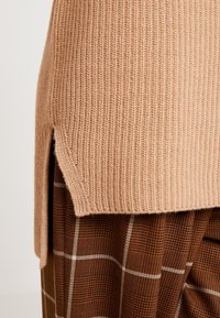 Beige ribbet striktrøje med sideslids, lagd over et brunt ternet tøj med hvide linjer og en løs pasform.