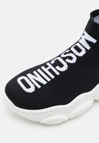 Černé slip-on tenisky s pletenou texturou, zdobené výrazným bílým textem a silnou bílou podrážkou. Zaoblená špička a flexibilní horní část.