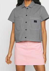 Camicia cropped a maniche corte con motivo pied de poule in bianco e nero, chiusura con bottoni, abbinata a una minigonna di denim rosa chiaro.