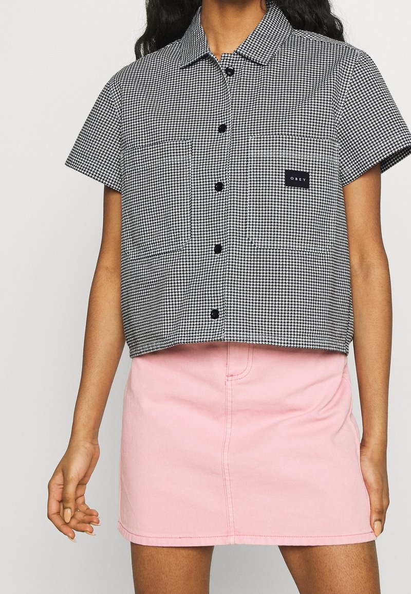 Camicia cropped a maniche corte con motivo pied de poule in bianco e nero, chiusura con bottoni, abbinata a una minigonna di denim rosa chiaro.