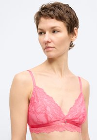 Γυναίκα με κοντά καστανά μαλλιά που φοράει ροζ δαντελωτό μπραλέτο με λεπτές τιράντες, που κοιτάζει ελαφρώς προς τα αριστερά της, με λευκό φόντο.