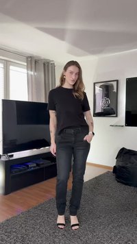 Sort t-skjorte med korte ermer kombinert med smale svarte jeans og svarte sandaler med hæl. Tilbehør inkluderer store gulløreringer. Teppebelagt gulv.