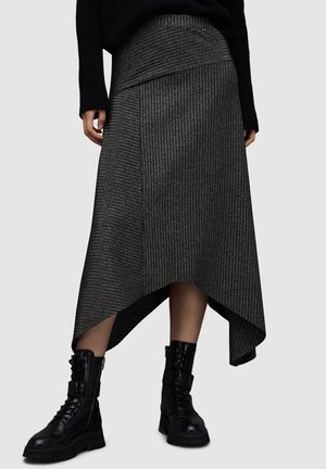 Femme portant une jupe asymétrique côtelée gris foncé et des bottes de combat noires à lacets, debout devant un fond clair uni.