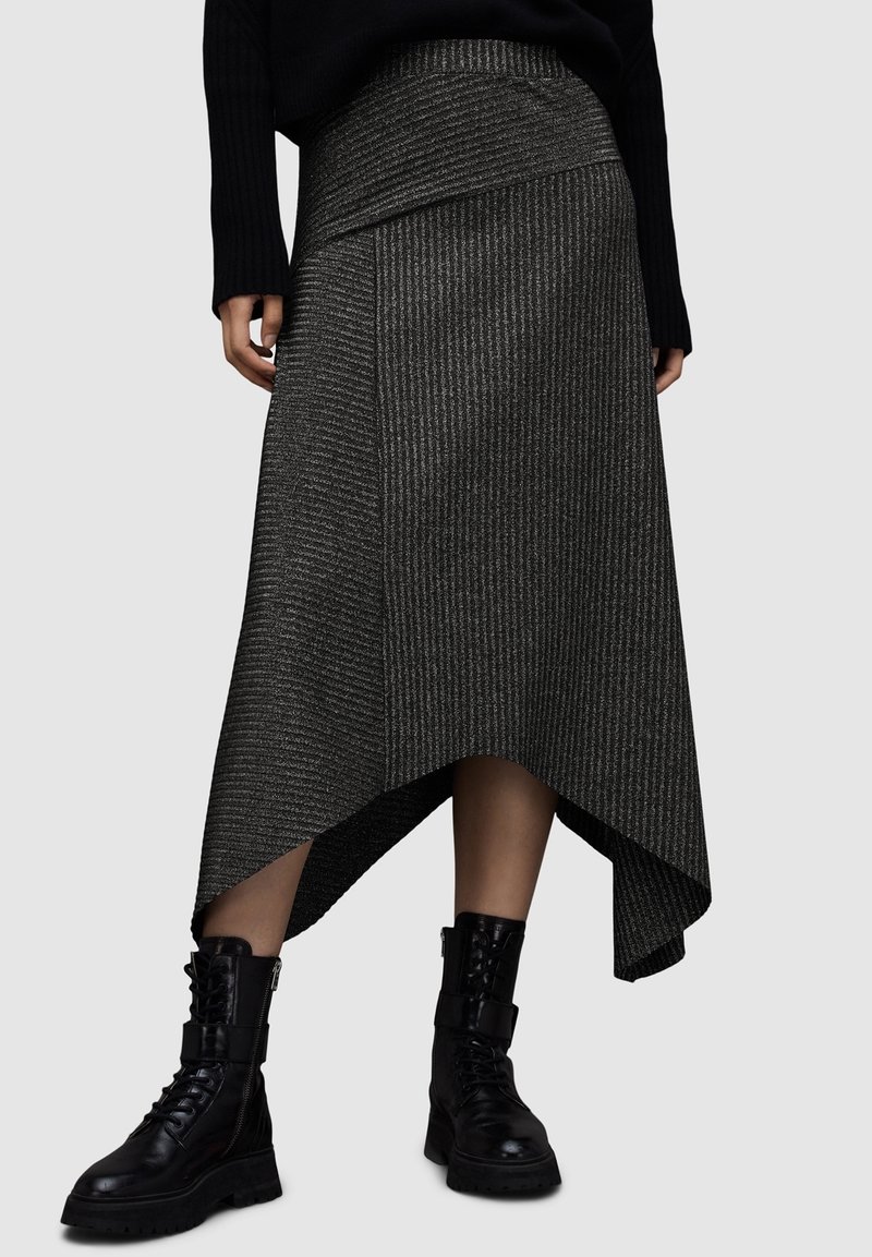 Femme portant une jupe asymétrique côtelée gris foncé et des bottes de combat noires à lacets, debout devant un fond clair uni.