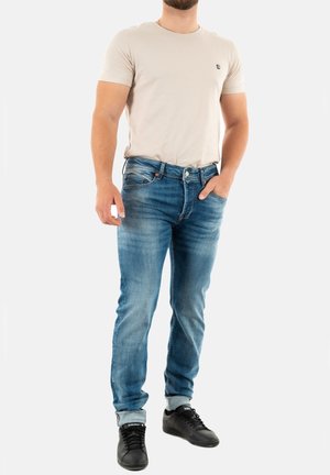 Camiseta de manga corta beige con un pequeño logo, combinada con unos vaqueros de denim azules con acentos desgastados y zapatillas negras.