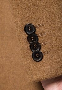 Brązowy wełniany rękaw kurtki z czterema czarnymi guzikami o gładkim wykończeniu i widocznym ściegniem. Teksturowany materiał o miękkim wyglądzie.