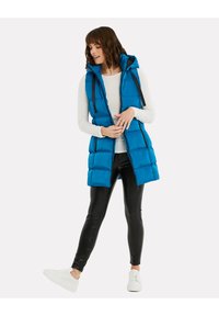 Blaues Pufferweste mit Kapuze, ausgestattet mit gesteppten Abschnitten und schwarzen Reißverschlussdetails. Kombiniert mit schwarzen Leggings und weißen Turnschuhen.