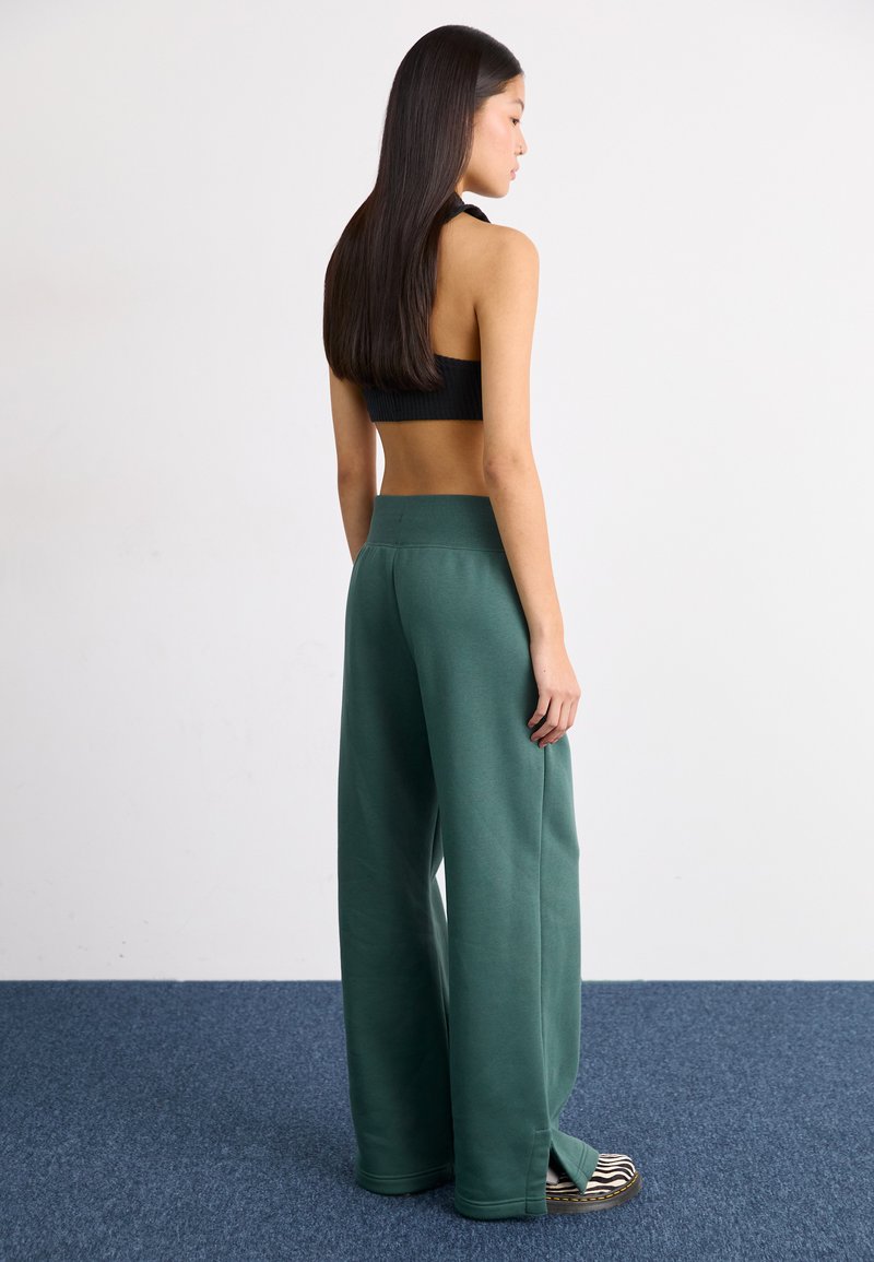 Dunkelgrüne, weit geschnittene Sweatpants mit hohem Bund, aus weichem Stoff. Das Modell kombiniert sie mit schwarz-weißen gestreiften Schuhen.