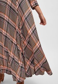 Jupe à carreaux flottante en rose, orange, noir et blanc, portée avec des bottes marron jusqu'aux genoux, avec une vue partielle d'un bras et d'une main de femme.