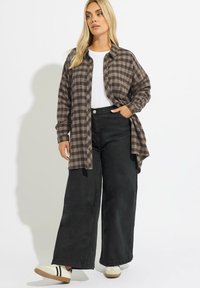 Camicia a quadri oversize in marrone scuro e beige sopra una maglietta bianca, abbinata a pantaloni a gamba larga neri e scarpe bianche con strisce nere.