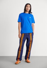 Blå bomulls-T-skjorte med en liten hvit logo, kombinert med marineblå bukser med oransje vertikale striper og et teksturert mønster. Brune treningssko.