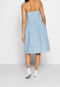 Ljusblå denimklänning med en åtsittande bodice, utsvängd kjol och tunna axelband. Har en rynkad midja och en liten logodetalj på baksidan.