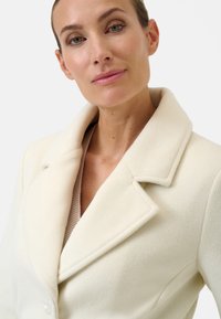 Cream-colored wool coat with a shawl collar and button closure. Soft texture with a structured design. Layered over a light sweater.