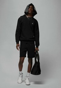 Felpa nera con piccolo logo, pantaloncini coordinati, scarpe da ginnastica bianche e borsa nera. La modella indossa un bandana fantasia come cappuccio.