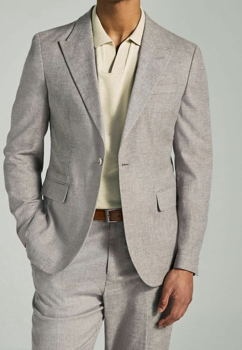 Homme portant un costume gris clair avec un polo beige et une ceinture marron, debout, une main dans la poche, sur un fond neutre.