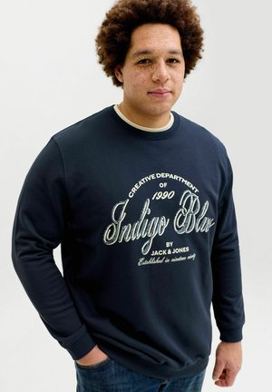 Ung mann med krøllete hår iført en marineblå genser med hvit tekst som sier "Creative Department of 1990 Indigo Blue by Jack & Jones."